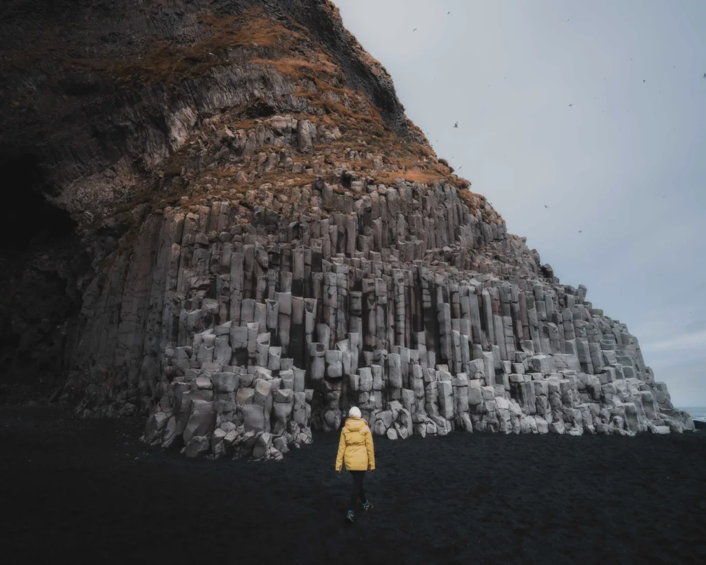 Woman in a yellow mackintosh stands in front of the impressive basalt rock formations on the black sand beach Reynisfjara in Iceland