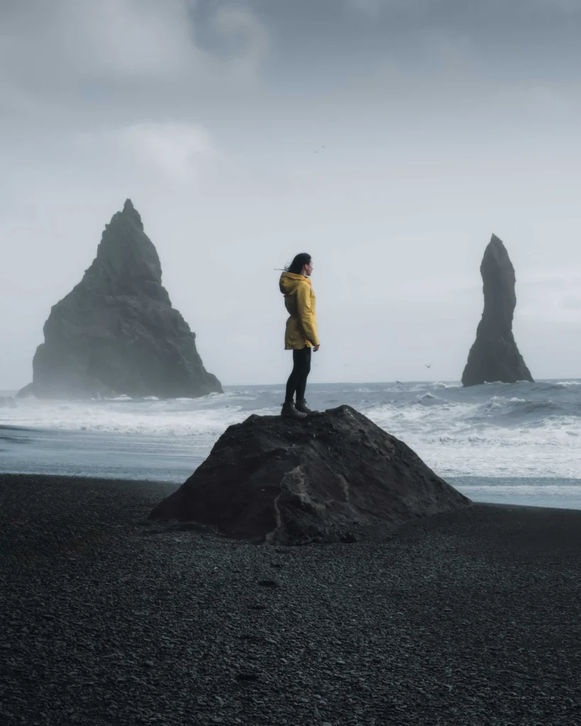 girl in yellow raingear stands on a hill at the beach reynisfjara in atmospheric scenery