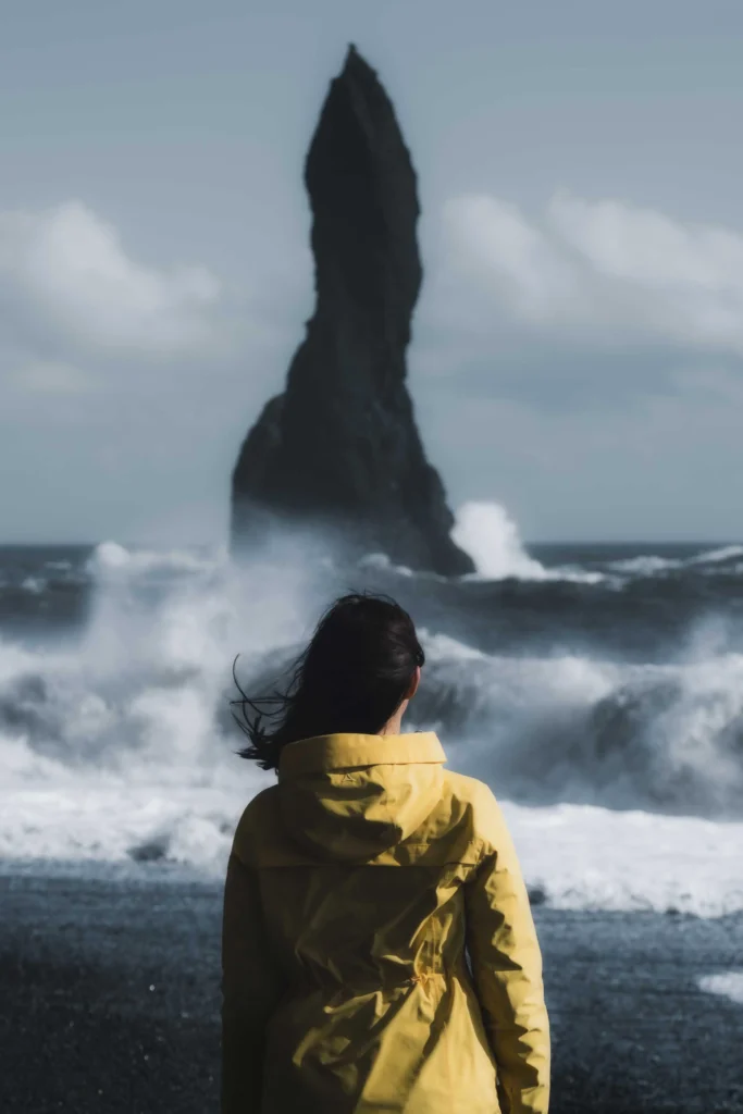 A woman in a yellow mackintosh walks along the black beach reynisfjara towards the rocks in front of the beach, which look like petrified trolls