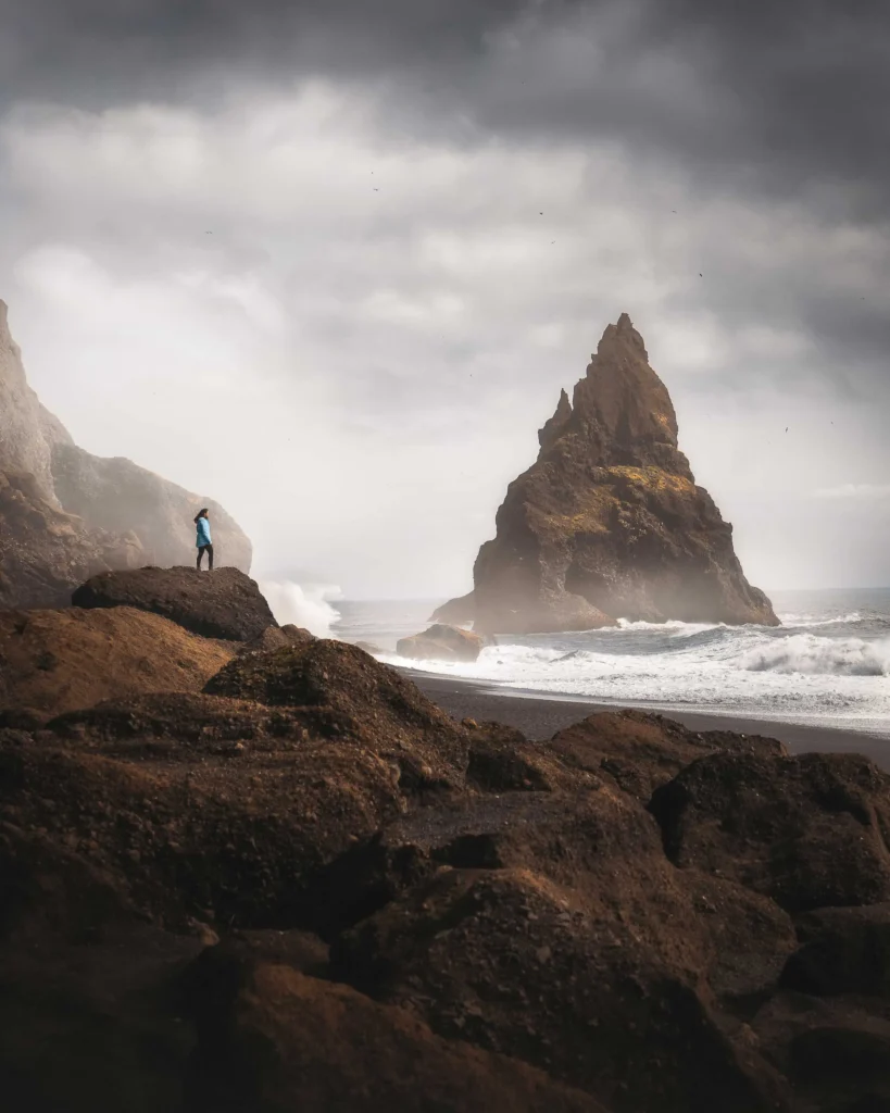 A woman stands on a rock on the black sandy beach of Reynisfjara and looks out over the rough North Atlantic Ocean, where rocks stand like petrified trolls